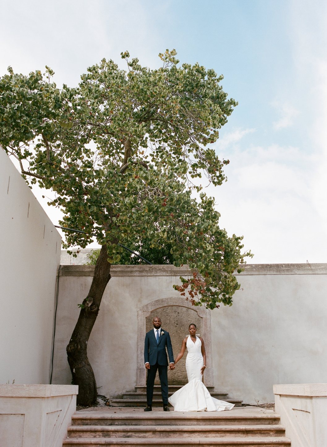 Mo & Toba holding hands on stone steps beneath a leaning tree, Lisbon